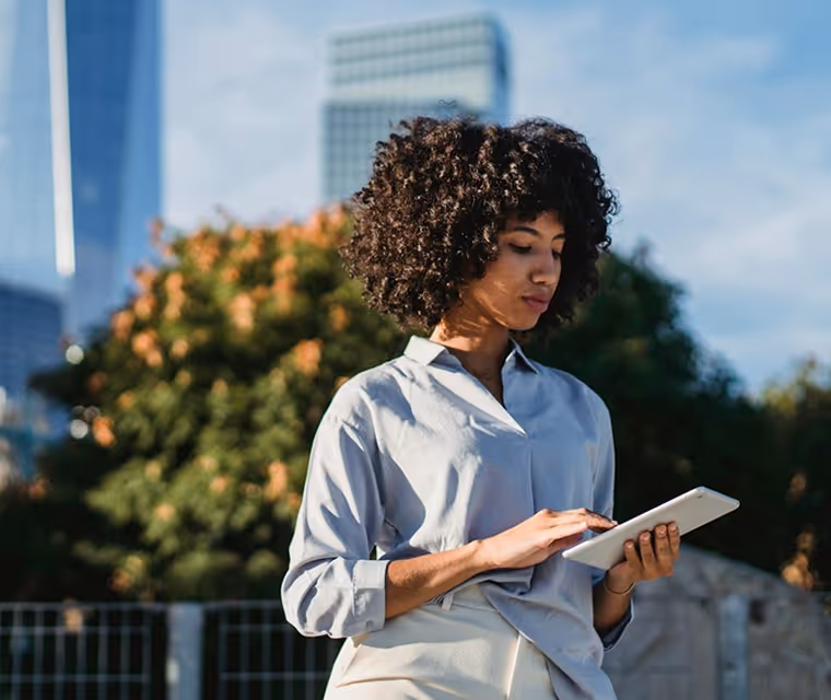Woman with curly hair standing outdoors using a tablet with trees and buildings in the background.