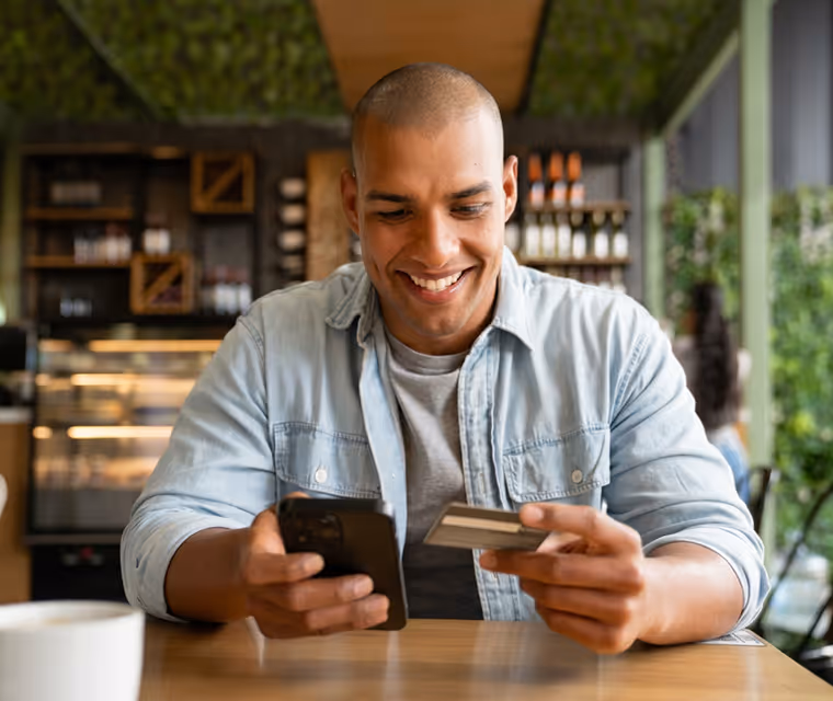 Smiling man in denim shirt holding a smartphone and credit card at a cafe table.