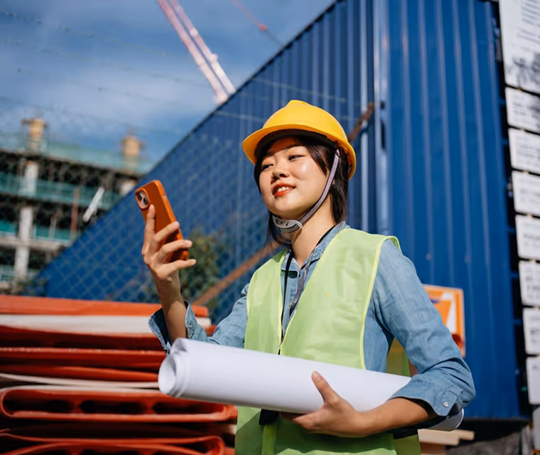 Construction worker in yellow helmet and vest holding blueprints and looking at a smartphone outdoors.