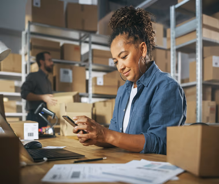 Woman using smartphone at a table in a warehouse surrounded by cardboard boxes and a laptop.