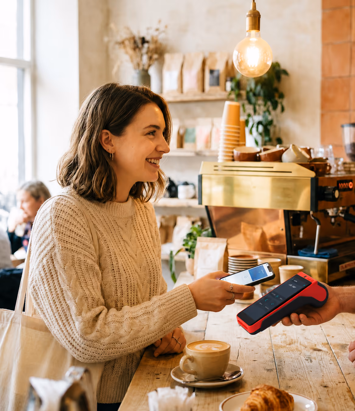 Woman smiling while paying with phone at a cafe counter with coffee and pastry nearby.