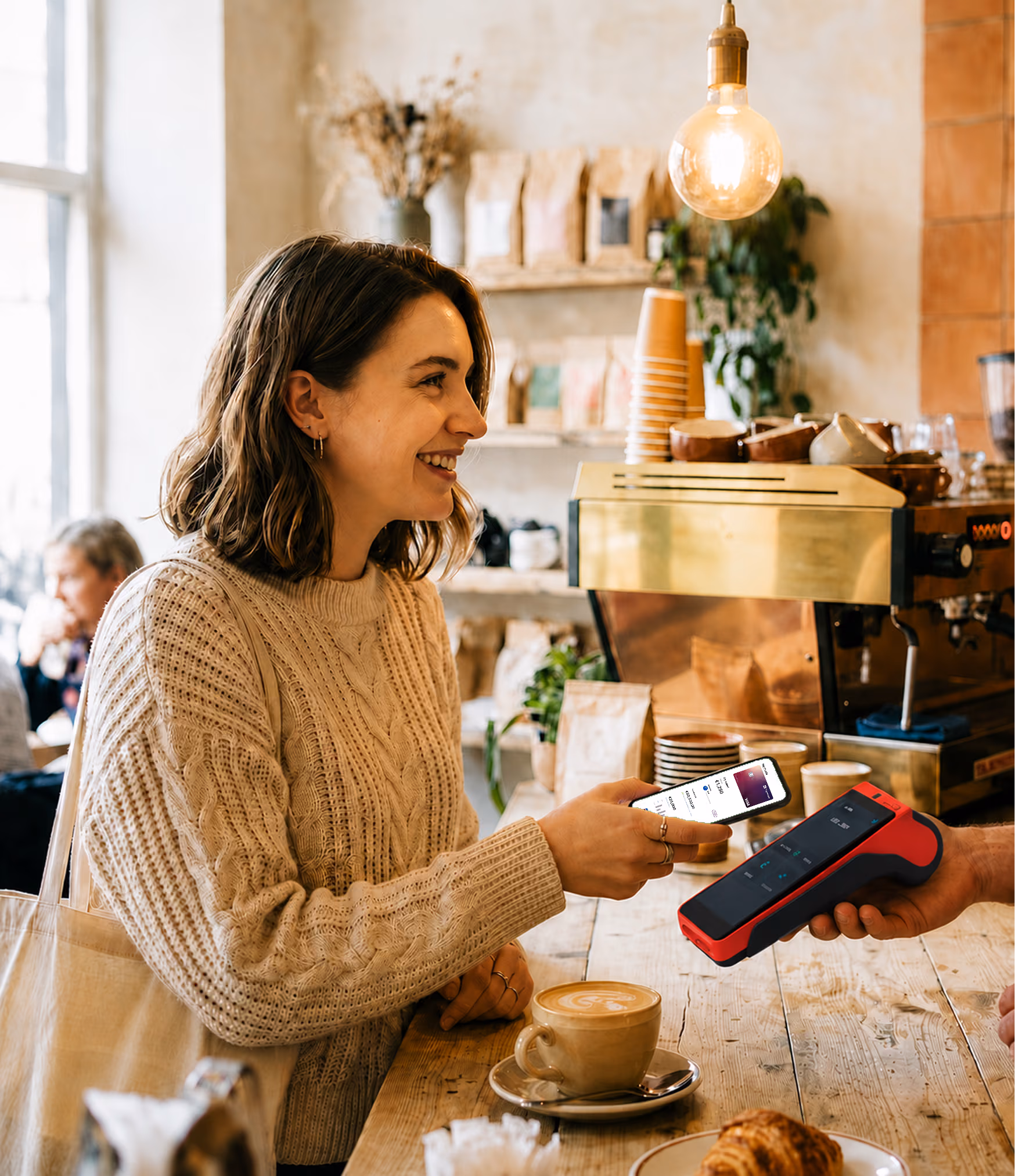 Woman smiling while paying with phone at a cafe counter with coffee and pastry nearby.