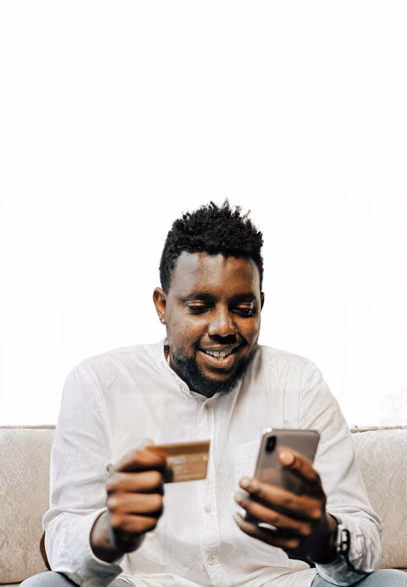 Man in white shirt smiling while holding a credit card and using a smartphone on a couch.