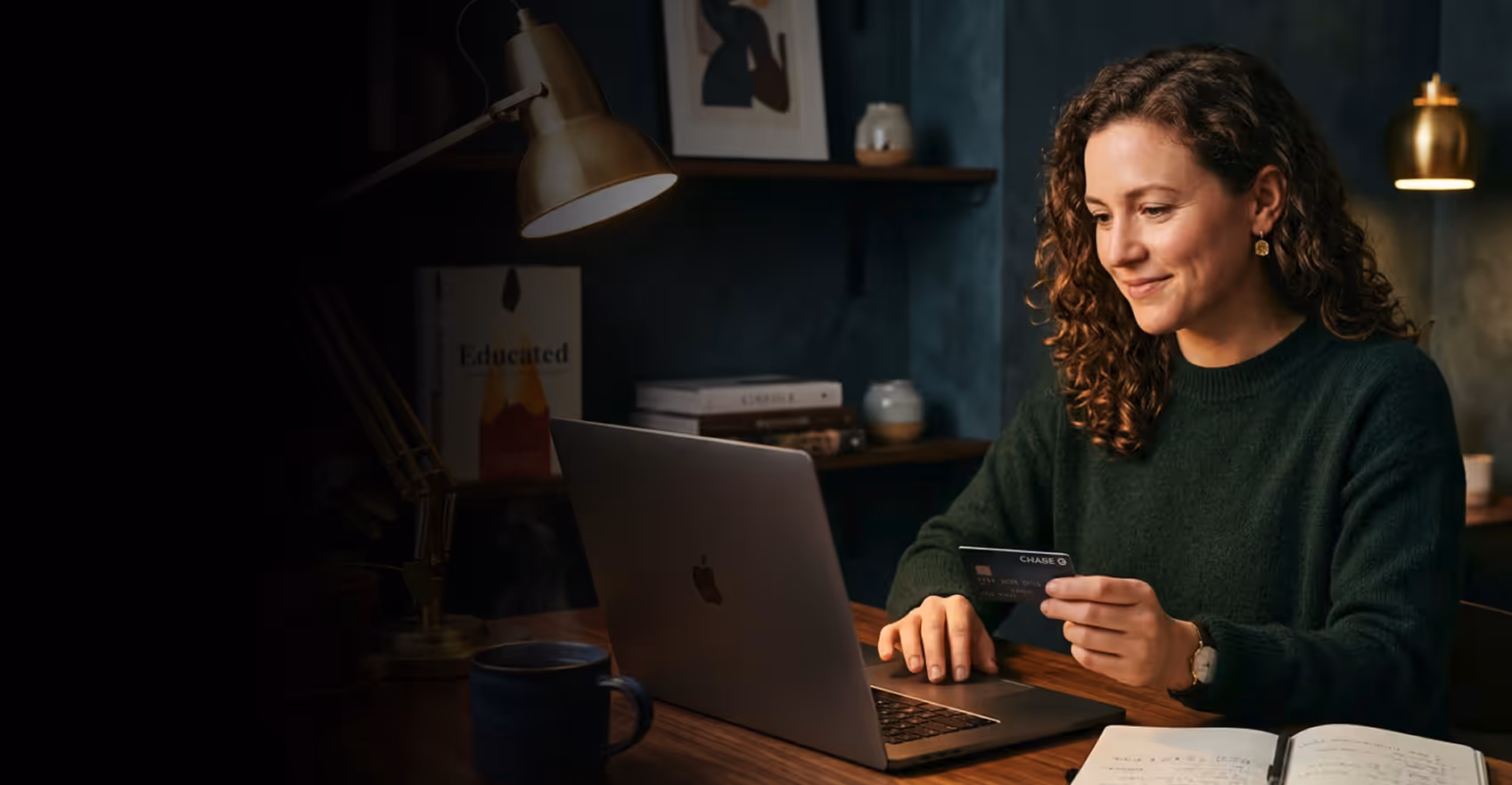 Woman sitting at desk using a laptop and holding a Chase credit card, smiling with open notebook nearby.