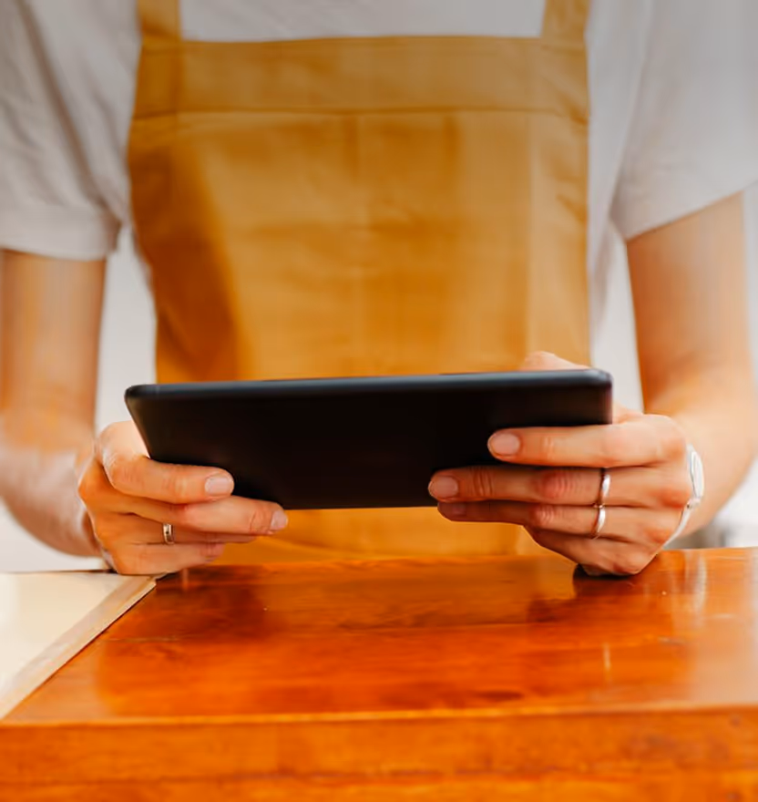 Person in tan apron holding a black tablet over a wooden counter.