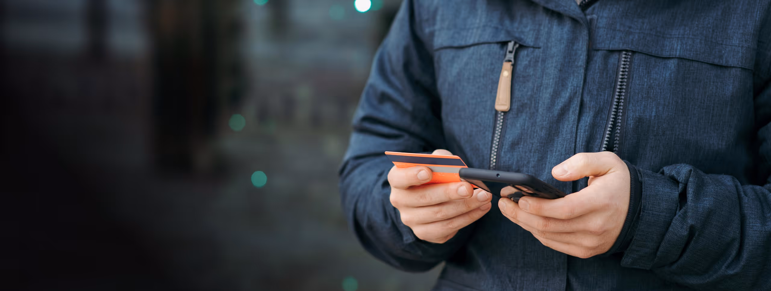 Person holding a credit card and using a smartphone, wearing a dark jacket with zippers.