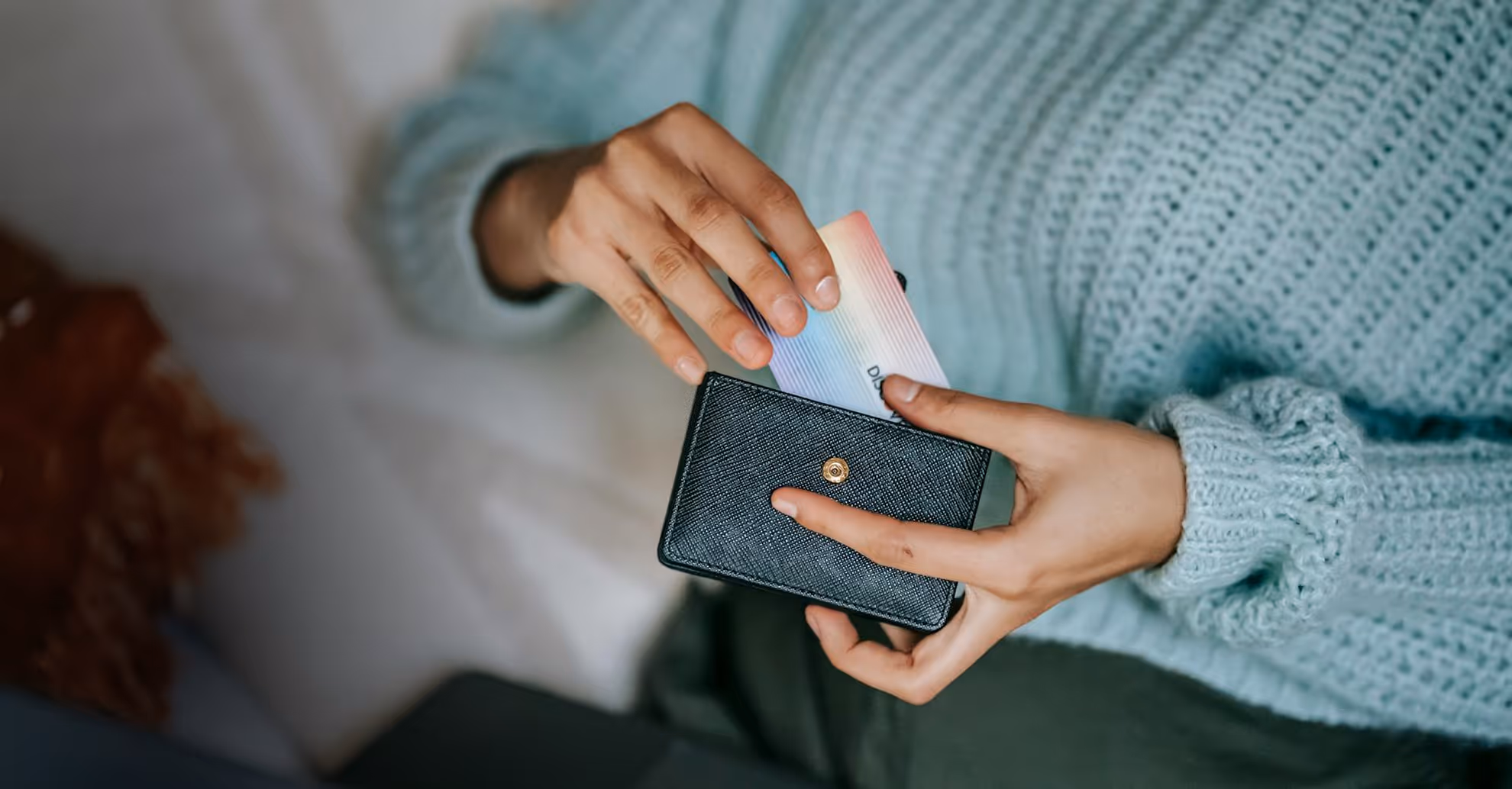 Person wearing a blue sweater putting a gradient-colored card into a black wallet.