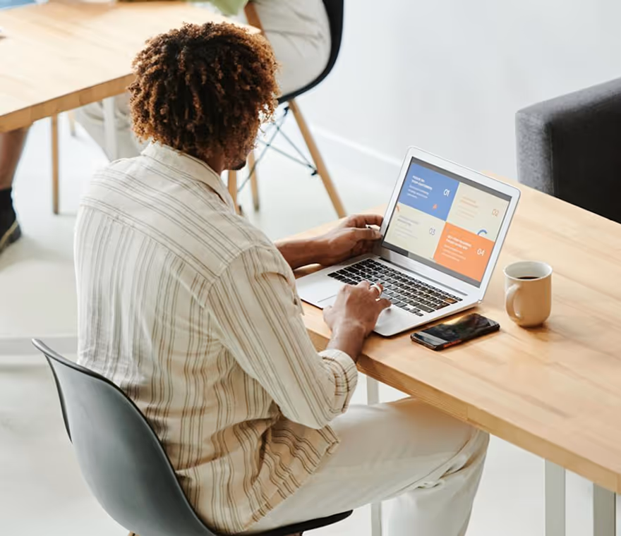 Person with curly hair working on a laptop at a wooden desk with a phone and coffee mug.