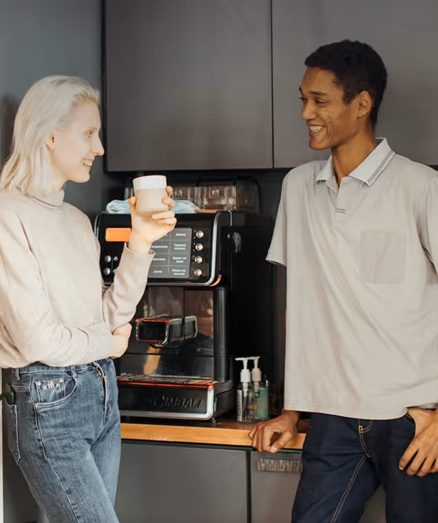 Two people smiling and chatting near a coffee machine, one holding a cup in a kitchen setting.