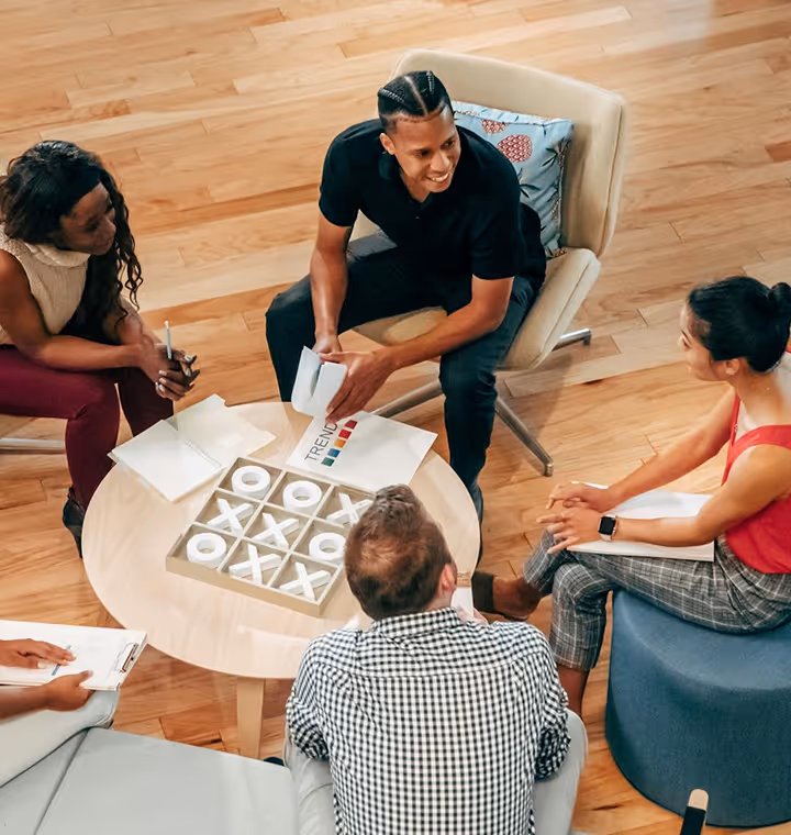 Four people gathered around a table playing tic-tac-toe and discussing with papers and notebooks.