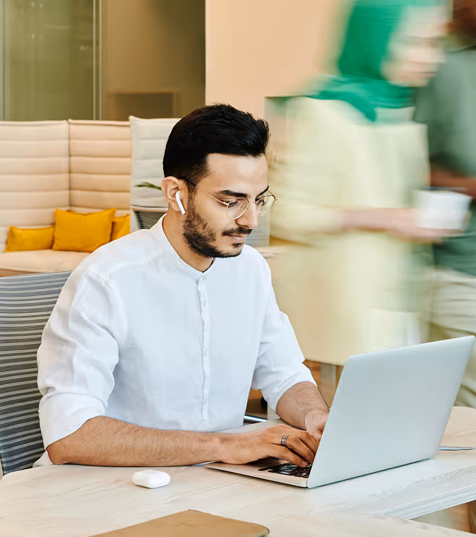 Man in glasses and earbuds working on a laptop at a desk in a modern office.