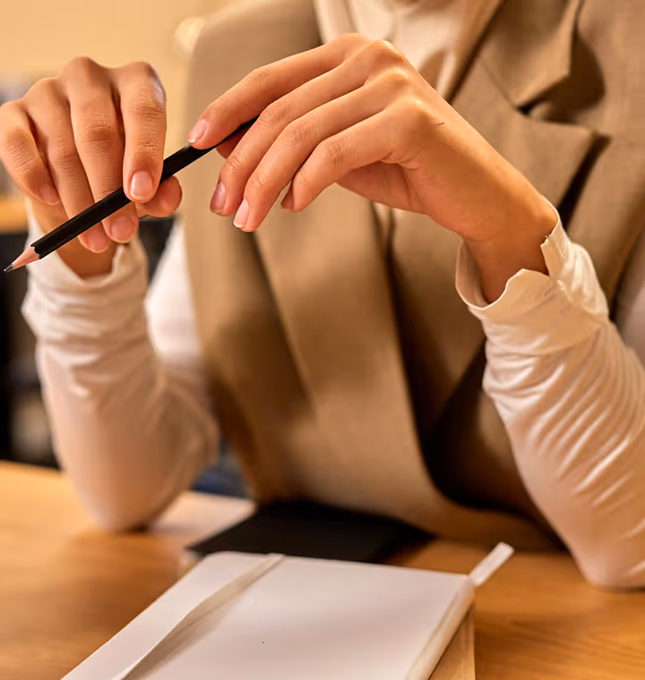 Person holding a black pencil above a closed white notebook on a wooden desk.
