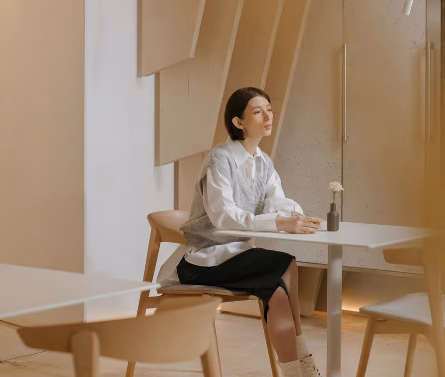 Woman sitting alone at a white table in a minimalist room with beige walls and wooden chairs.