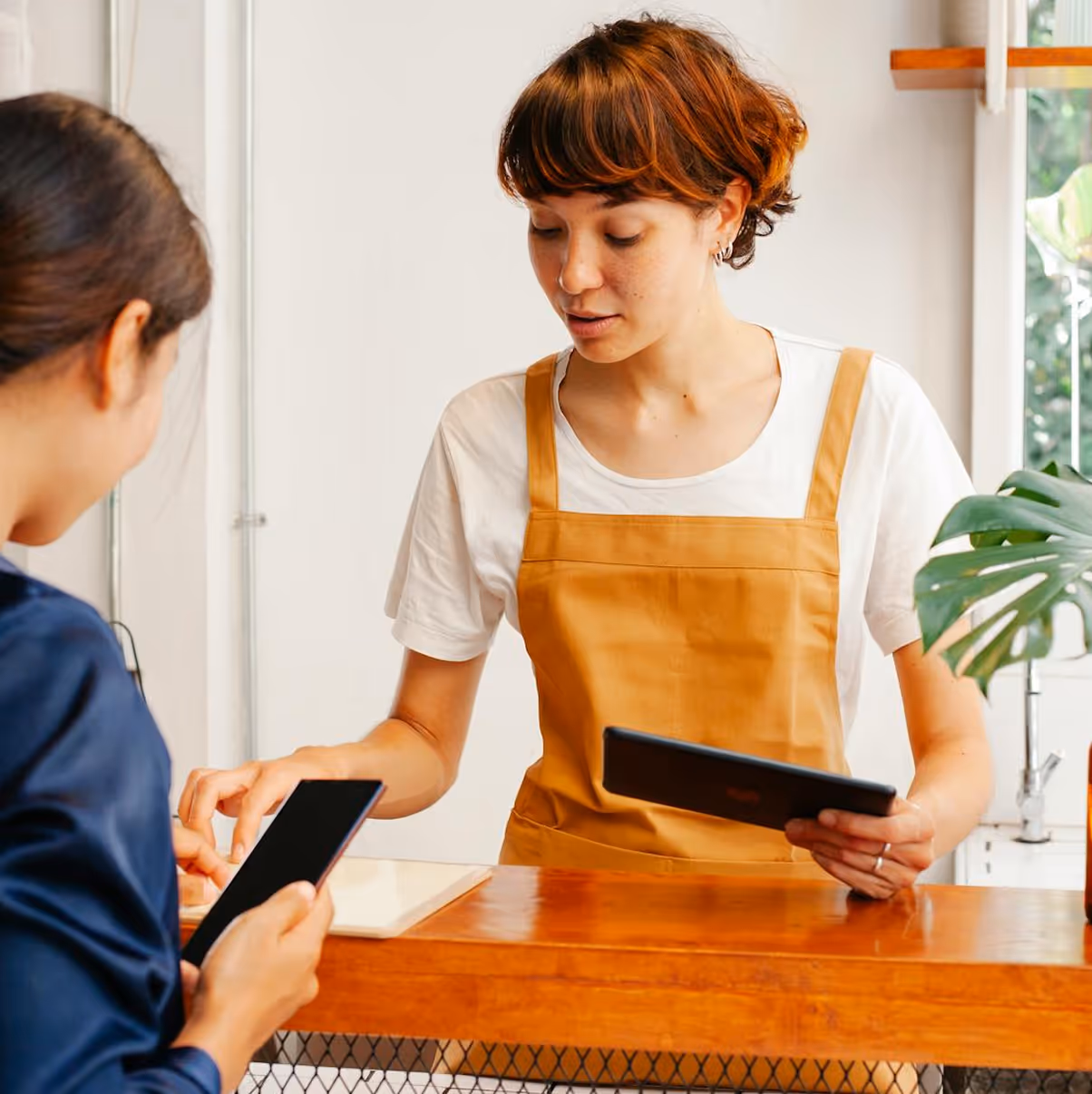 Woman in brown apron using a tablet at a wooden counter, serving a customer holding a smartphone.