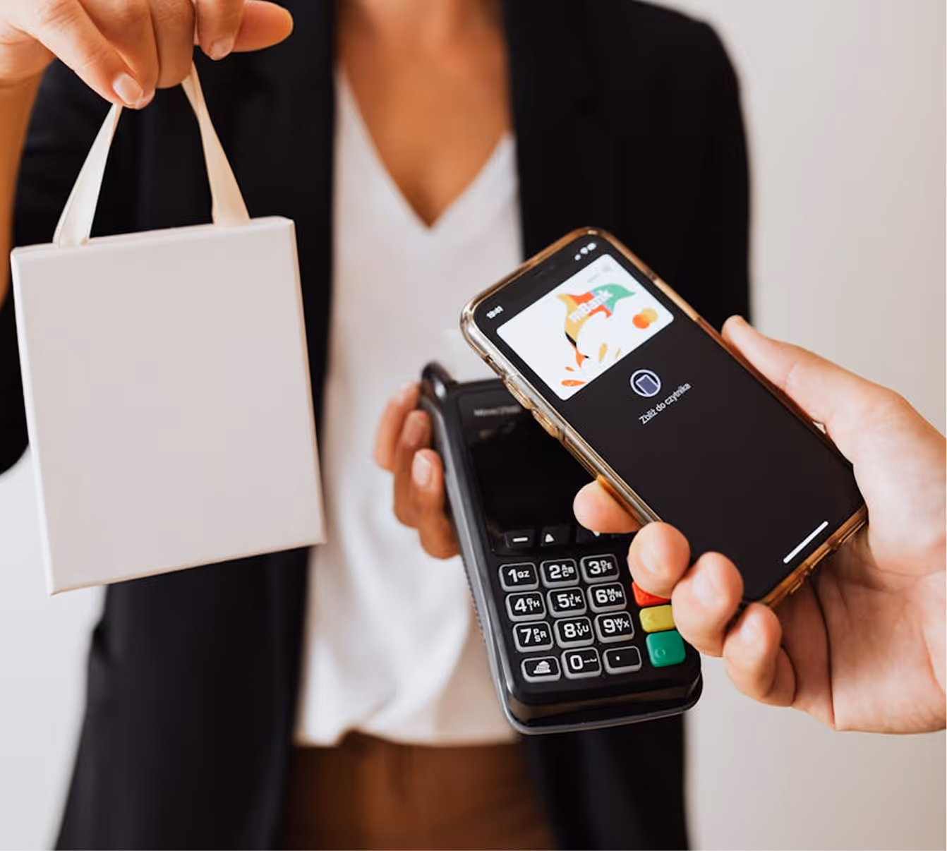 Person holding a white shopping bag and a card reader while another pays with a phone via contactless payment.