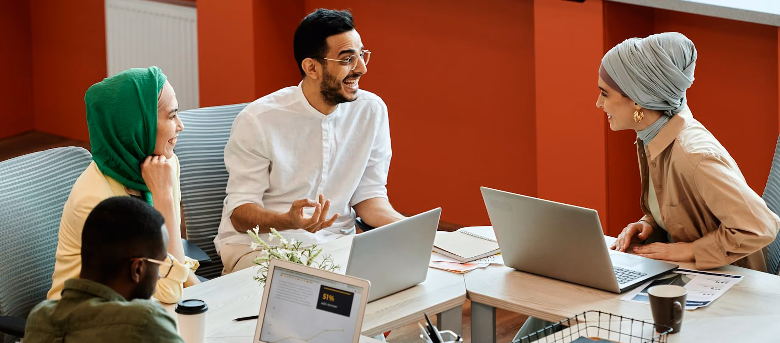 Four diverse colleagues laughing and talking while working with laptops in a modern office.