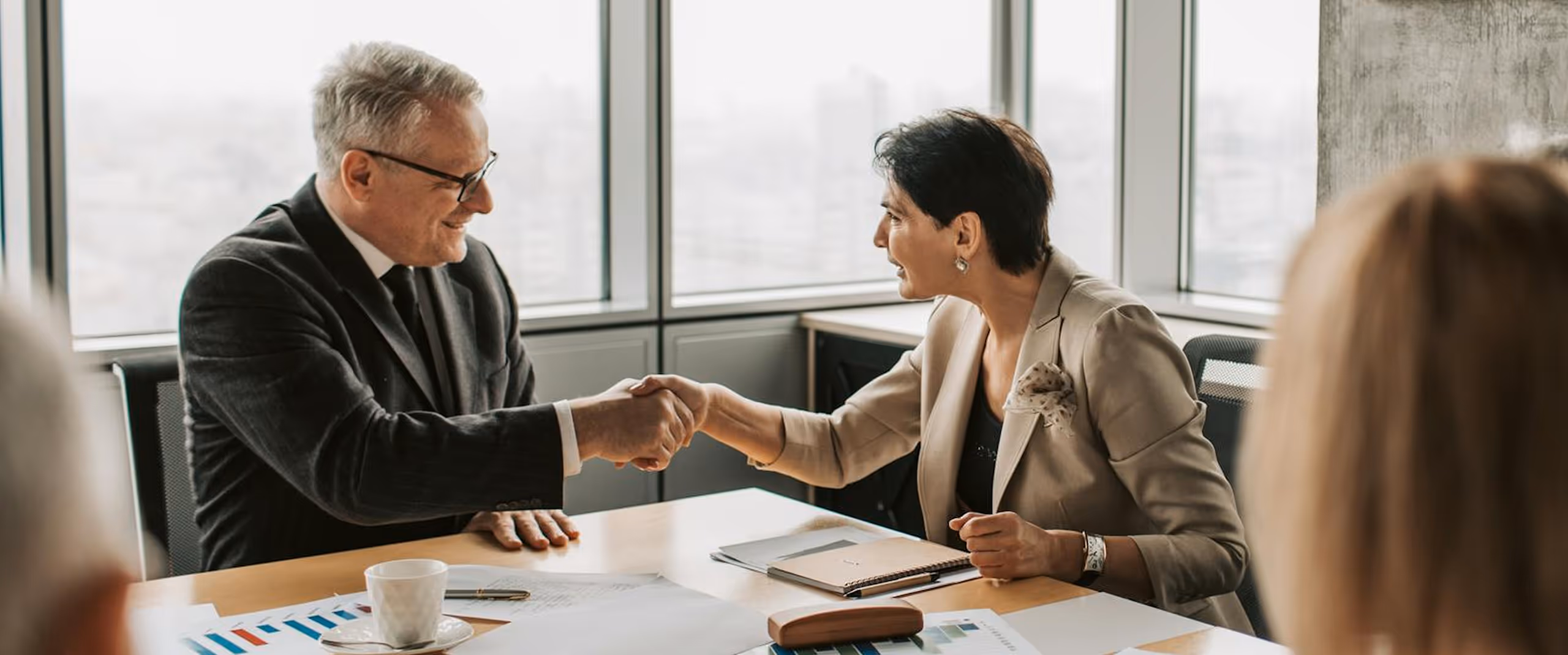 Two business professionals shaking hands across a desk with documents and coffee cup.