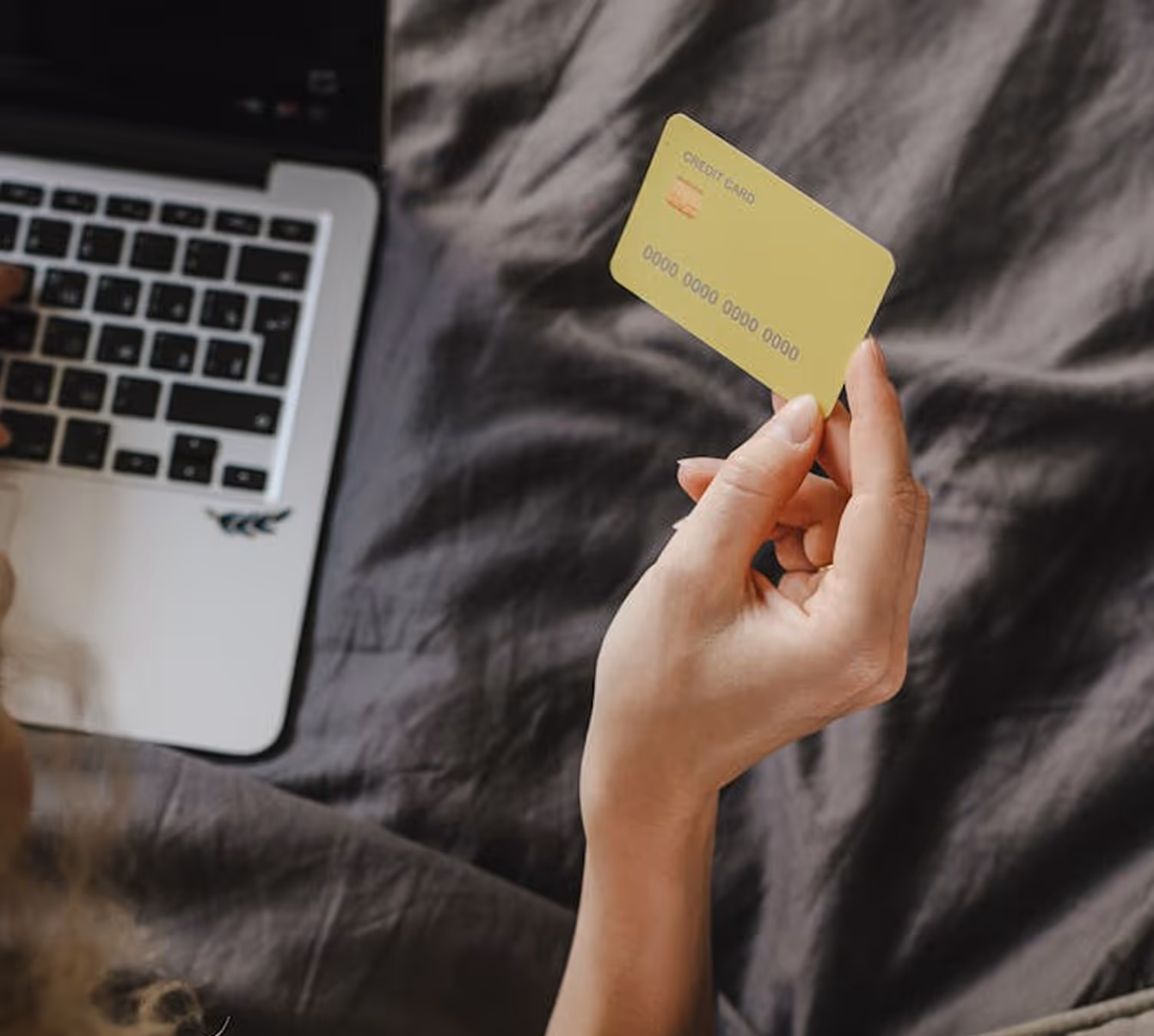 Person holding a yellow credit card near a laptop on a dark fabric surface.