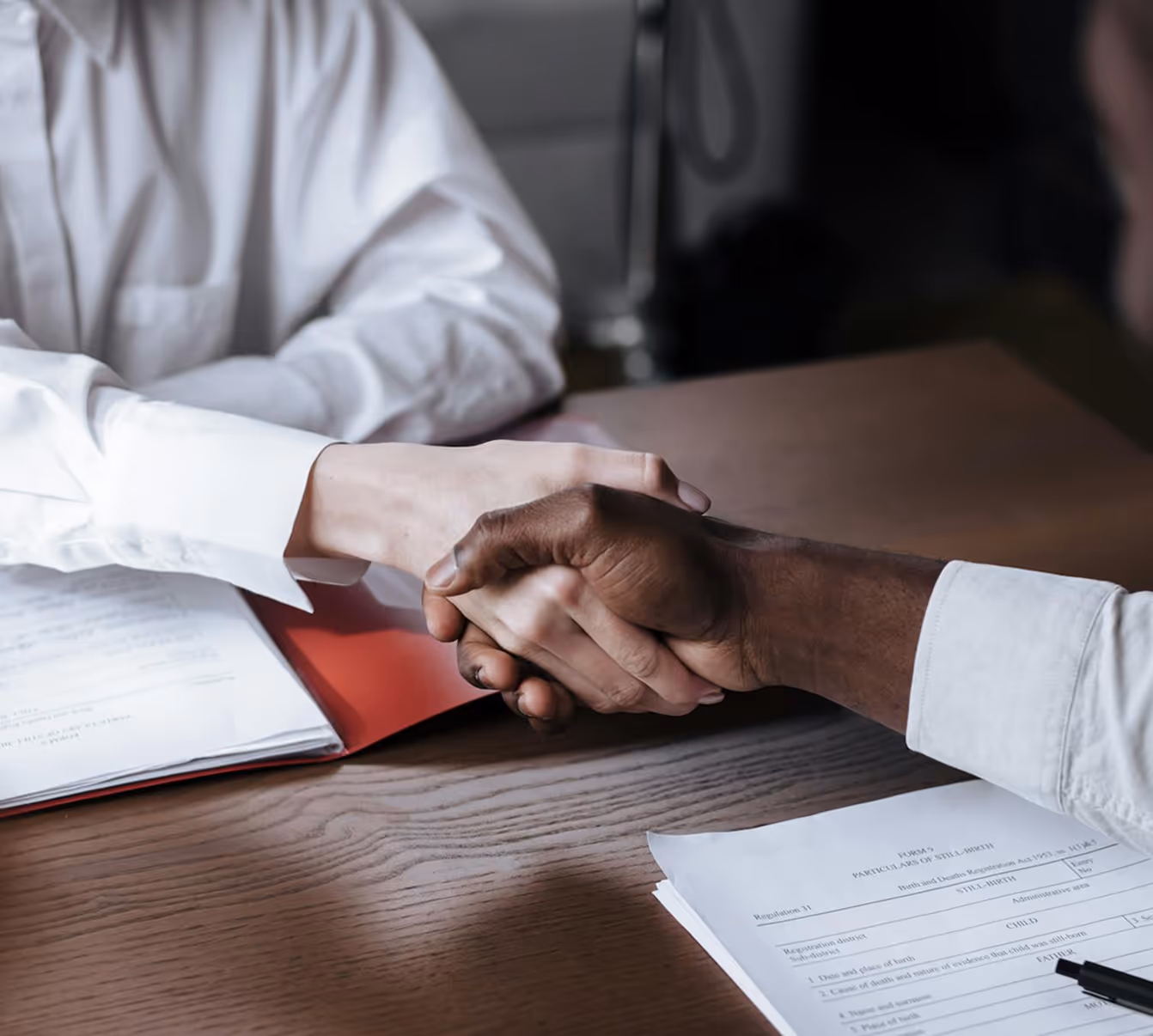 Two people shaking hands over a desk with documents and a pen.