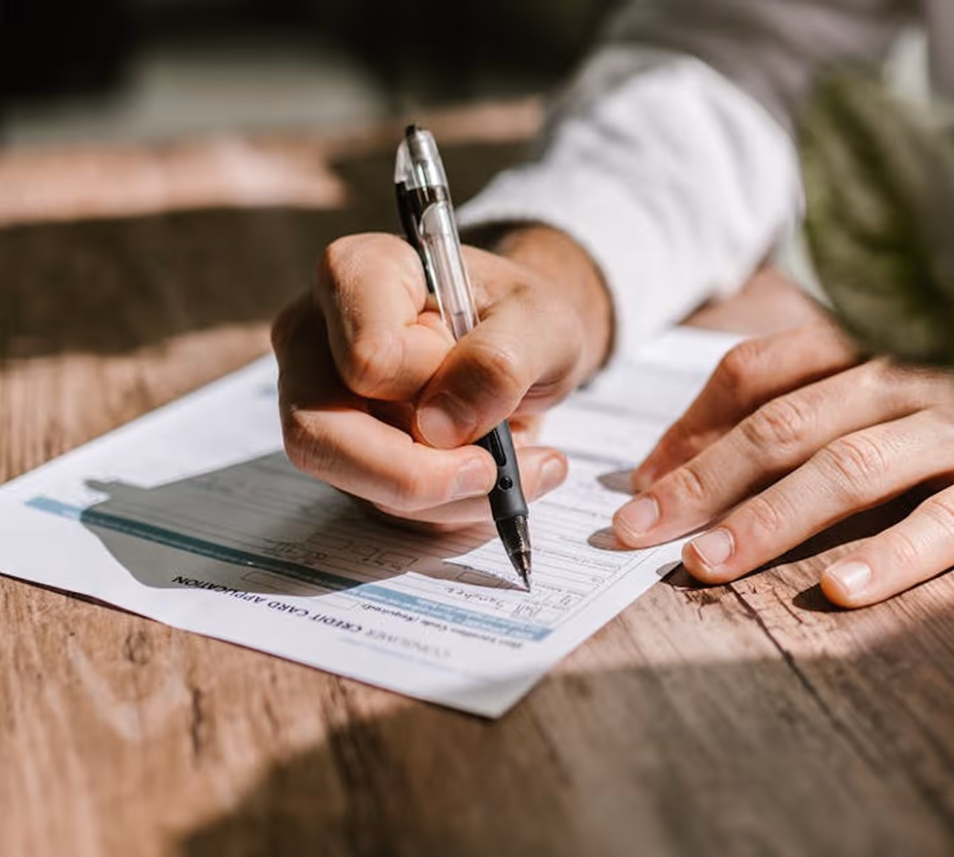 Close-up of a person filling out a credit card application form with a black pen on a wooden table.