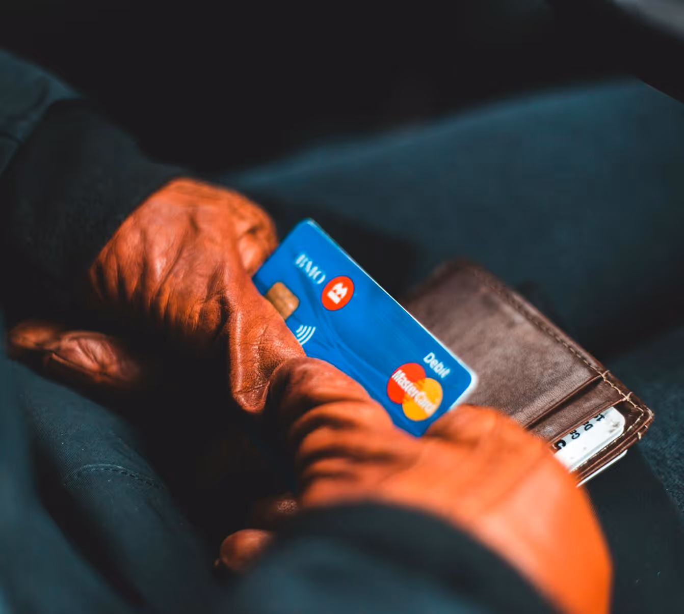 Person wearing brown gloves holding a blue debit Mastercard and a brown wallet.