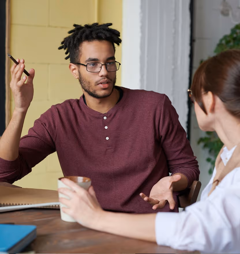 Two people having a discussion at a table, one holding a cup, the other gesturing with a pen.