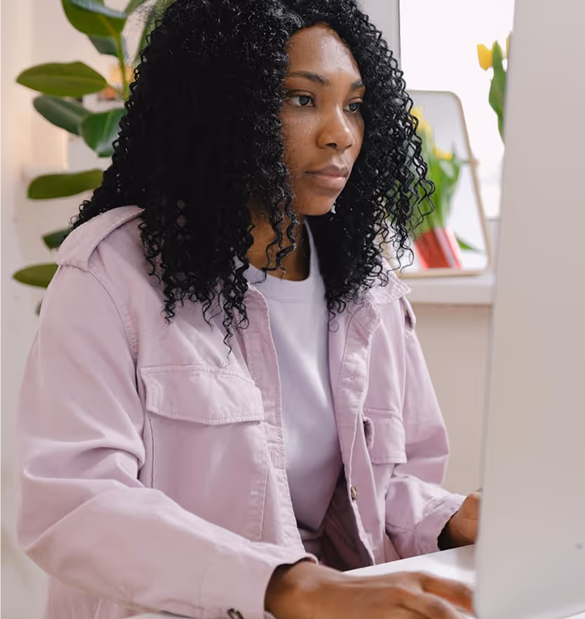 Focused woman with curly hair working at a computer desk with plants in the background.