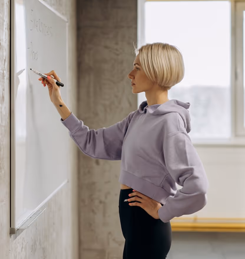 Woman in a lilac hoodie writing on a whiteboard in a bright room with large windows.