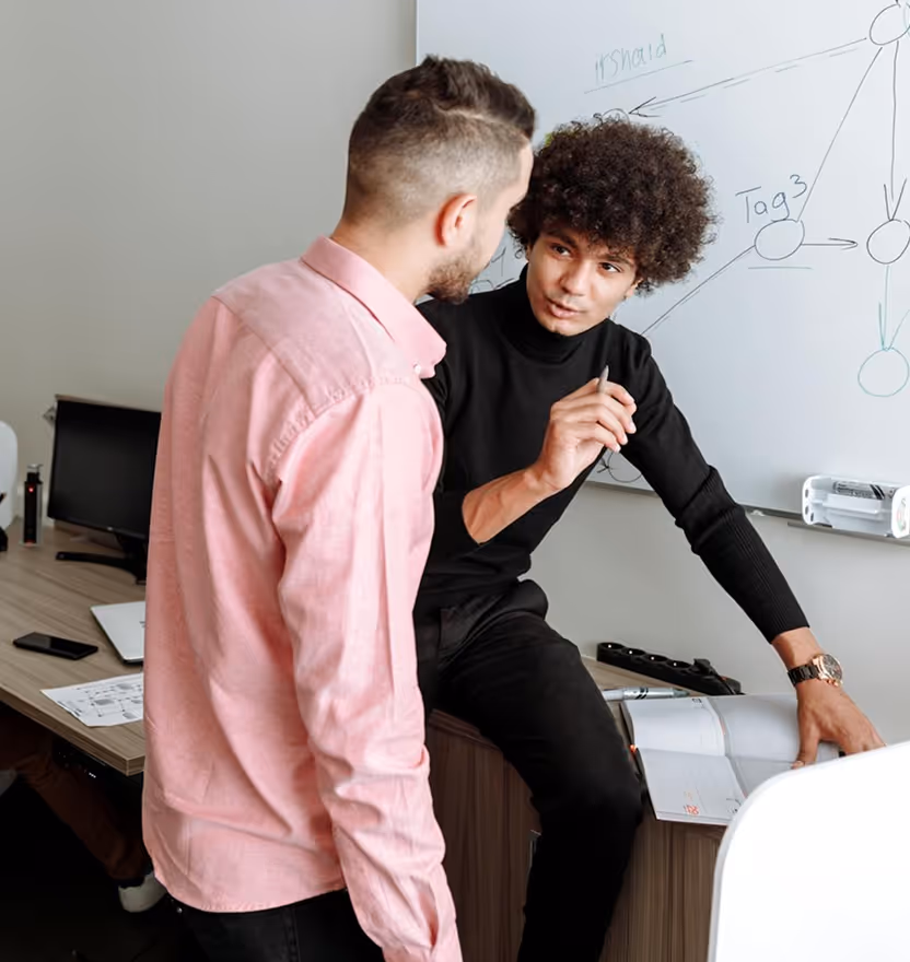 Two men discussing ideas by a whiteboard with diagrams in a modern office setting.