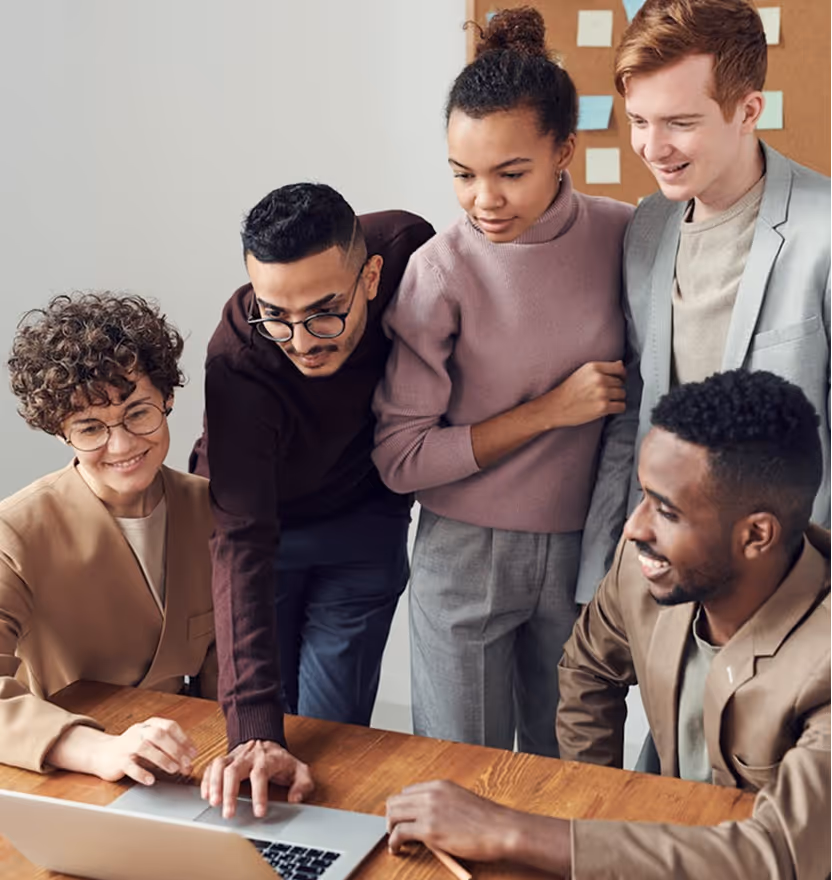 Five diverse professionals gathered around a laptop collaborating and smiling in an office setting.