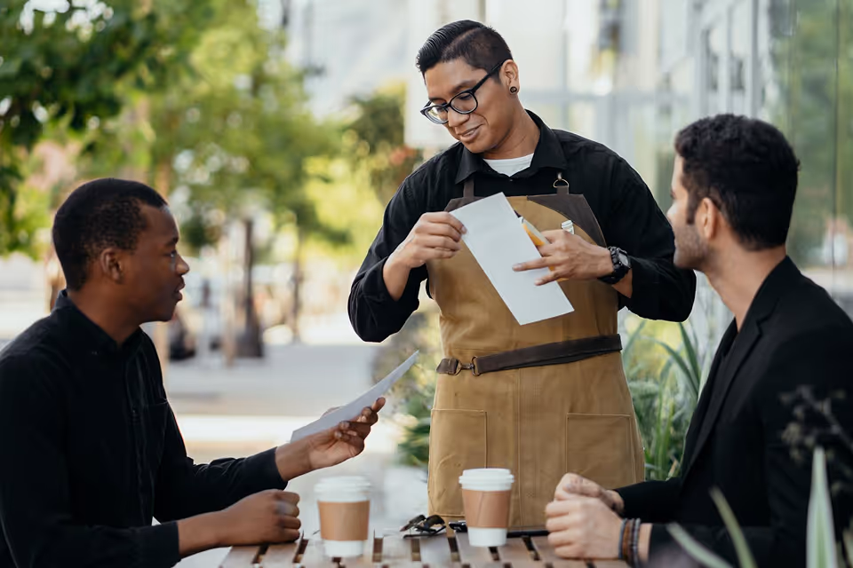 Barista in apron showing menu to two men seated at outdoor café table with coffee cups.