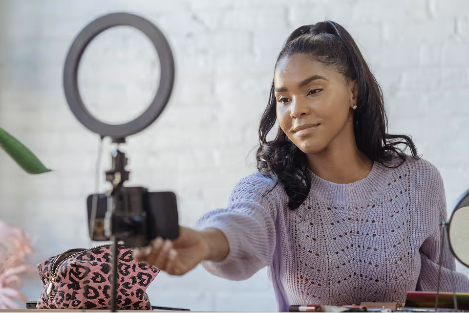 Woman in purple sweater recording herself with smartphone and ring light at makeup table.