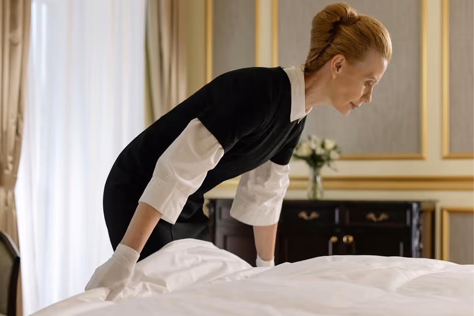 Hotel maid wearing white gloves making a bed in a well-decorated room with curtains and flowers.