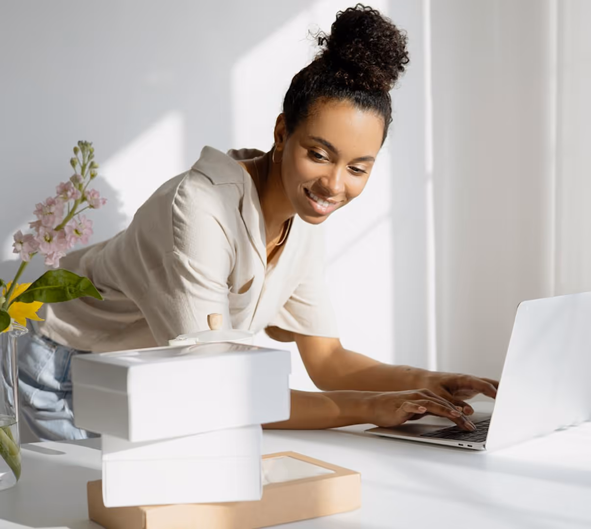 Smiling woman with curly hair typing on a laptop at a white desk with stacked boxes and flowers.