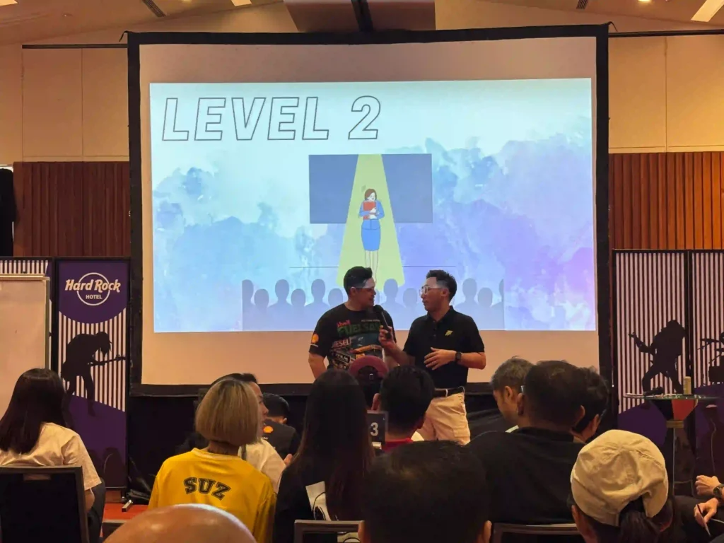 Two facilitators from Vision Building are on stage in front of a large screen displaying "LEVEL 2" and an illustration of a person in a spotlight. They are actively engaging the audience, who are seated and looking towards the stage and screen, illustrating a structured approach to learning and fostering a culture of enhanced perception beyond mere activities.