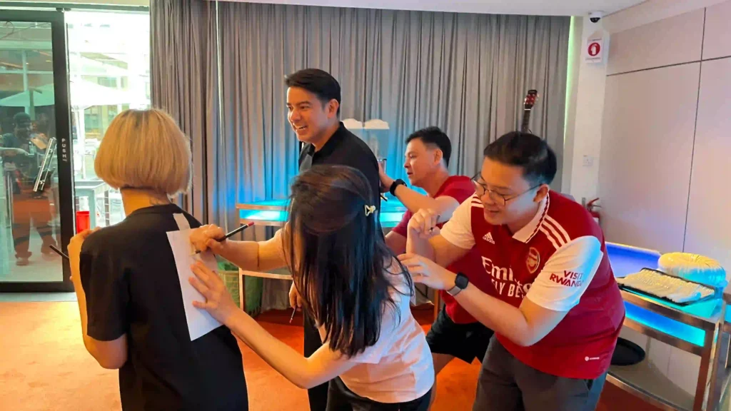 A group of five people from Vision Building are participating in a lighthearted team-building game where they are writing on papers taped to each other's backs. Their focused and amused expressions highlight the fun, collaborative play that can lead to deeper insights through reflection.