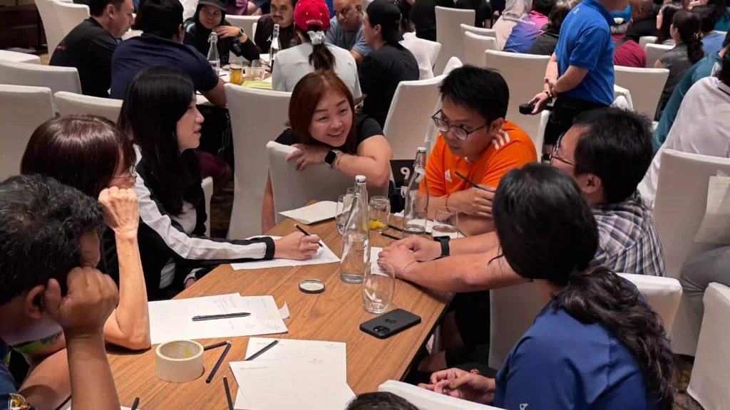customer of vision building are seated around a table, actively engaged in a focused discussion. They are taking notes on paper and using a variety of materials, illustrating a collaborative process of turning a shared purpose into actionable plans.