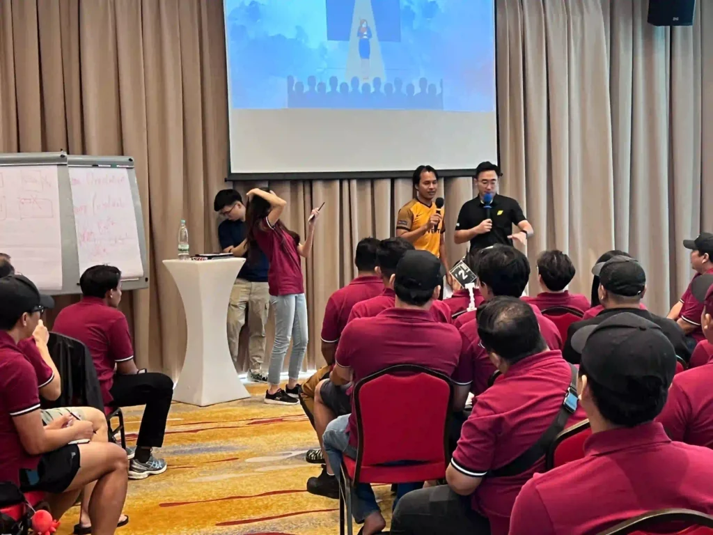 A group of professionals from Vision Building, wearing red polo shirts, are seated in an audience listening to two facilitators on stage. The facilitators are leading a session with a projector screen and a whiteboard in the background, representing the shift from a fun activity to a more focused evaluation and learning phase.