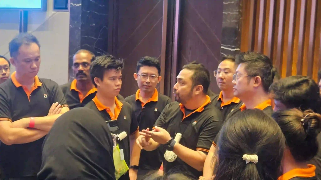 In Vision Building event,A diverse group of professionals wearing matching black and orange polo shirts are gathered in a circle, attentively listening to a man in the center who is speaking animatedly and gesturing with his hands. The setting appears to be an indoor corporate event or team-building session, emphasizing group discussion and interaction.
