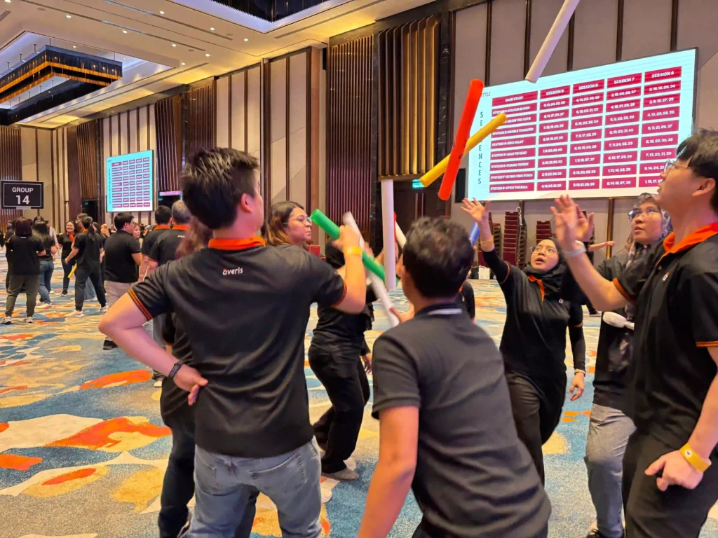 A group of professionals from Averis are playing a team-building game with colorful foam sticks in a large event hall, capturing a single moment of high-energy fun.