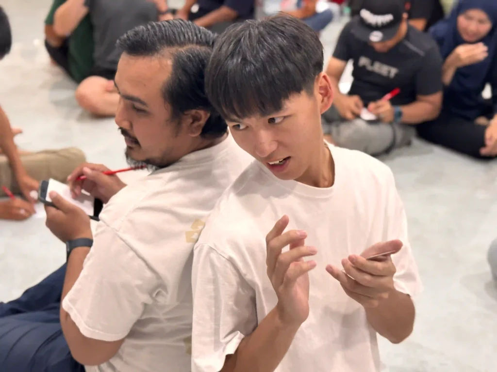 team members sit back-to-back, one writing and one gesturing in a group activity, symbolizing communication, perspective, and collaborative problem-solving in a team building exercise in malaysia