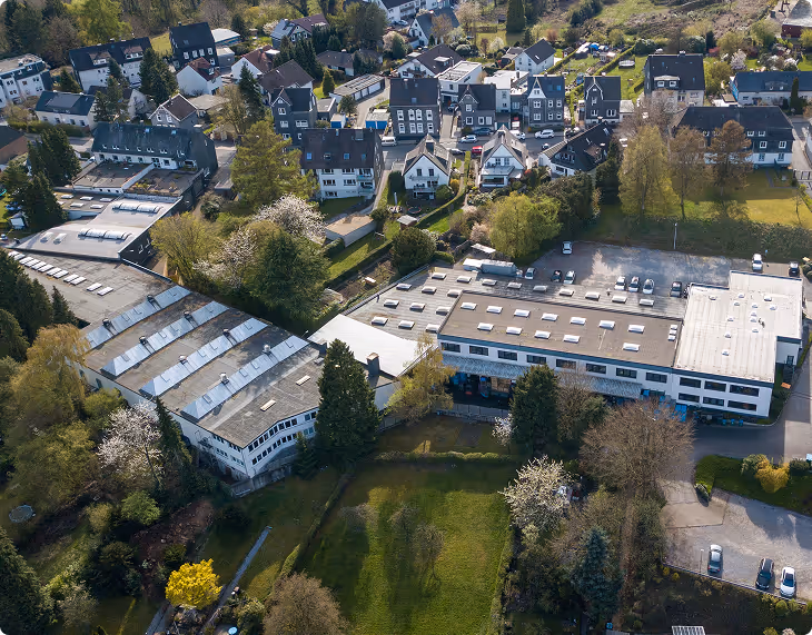 Luftaufnahme eines Industriegebiets mit mehreren Gebäuden und umgebenden Einfamilienhäusern in einer grünen, bewaldeten Gegend.