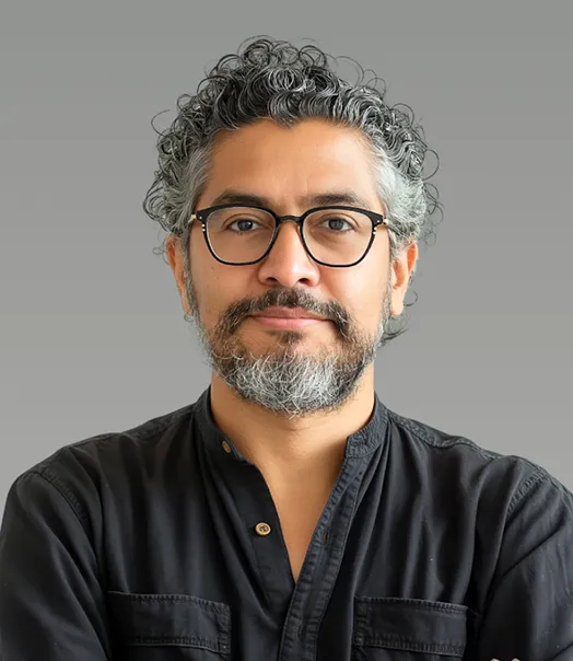 Man with curly gray hair and beard wearing glasses and a black shirt against a gray background.
