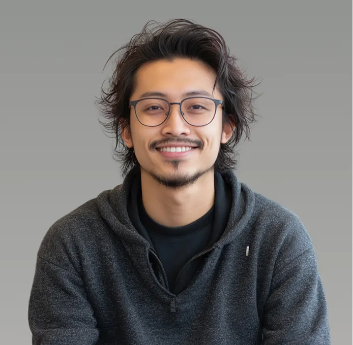 Smiling young man with glasses and dark hair wearing a dark gray hoodie against a gray background.