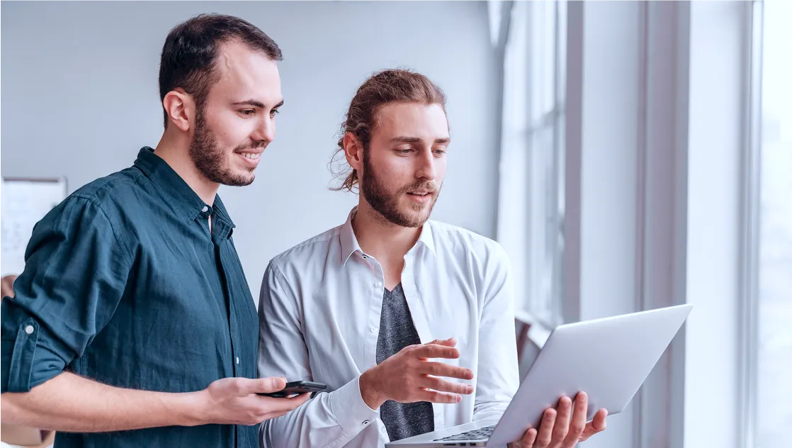 Two men standing indoors by a large window, one holding a laptop and explaining something to the other man holding a smartphone.