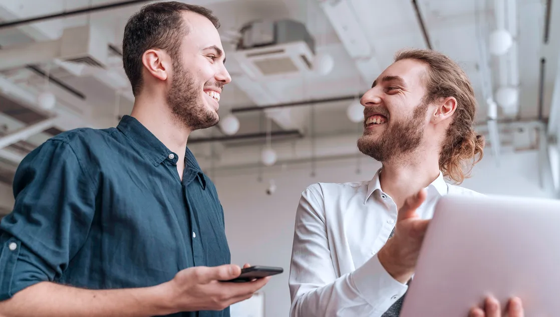 Two men smiling and conversing indoors, one holding a smartphone and the other a laptop.