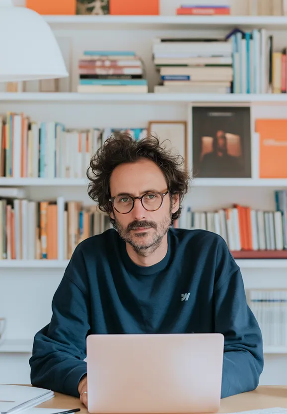 Man with curly hair and glasses sitting at a desk with a laptop in front of a bookshelf filled with books.
