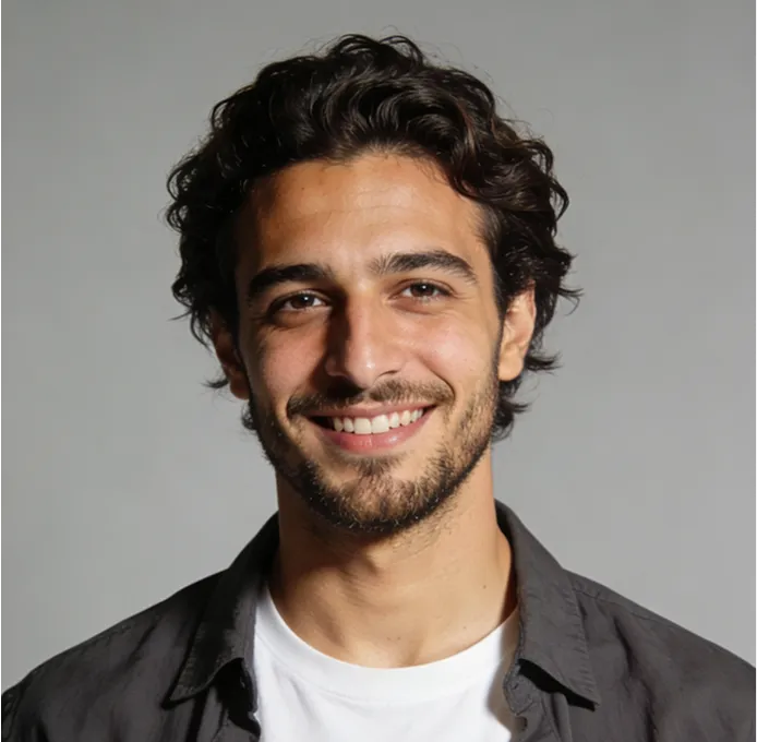 Smiling young man with dark curly hair and beard wearing a white shirt and dark jacket against a plain gray background.