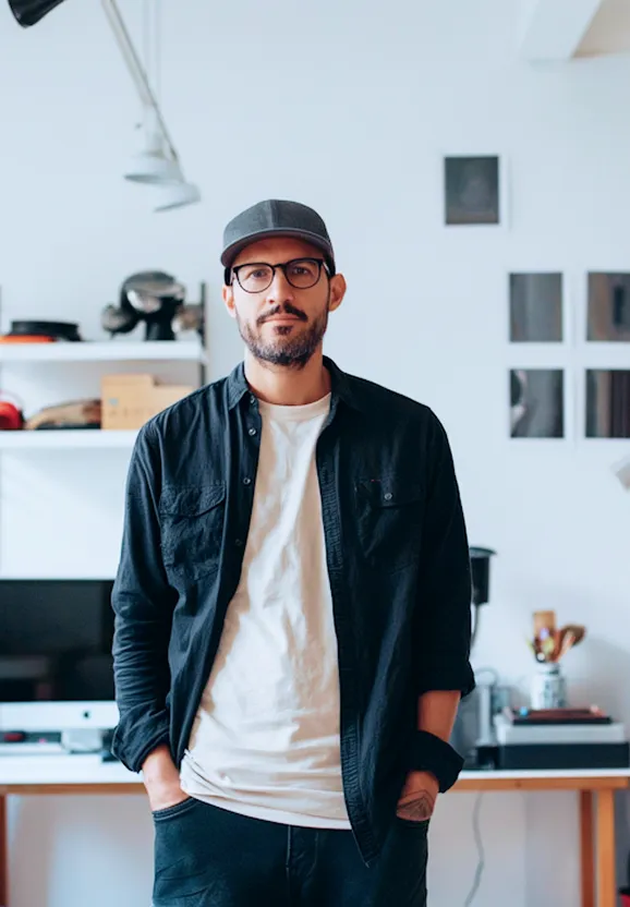 Man wearing glasses, a gray cap, white t-shirt, and black shirt standing in a modern office with hands in pockets.