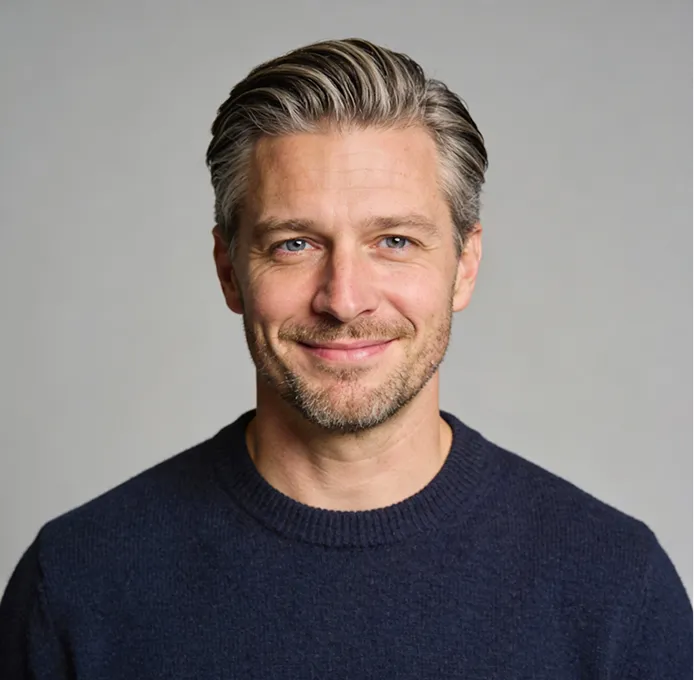 Smiling middle-aged man with short gray-streaked hair and stubble wearing a dark blue sweater against a plain gray background.